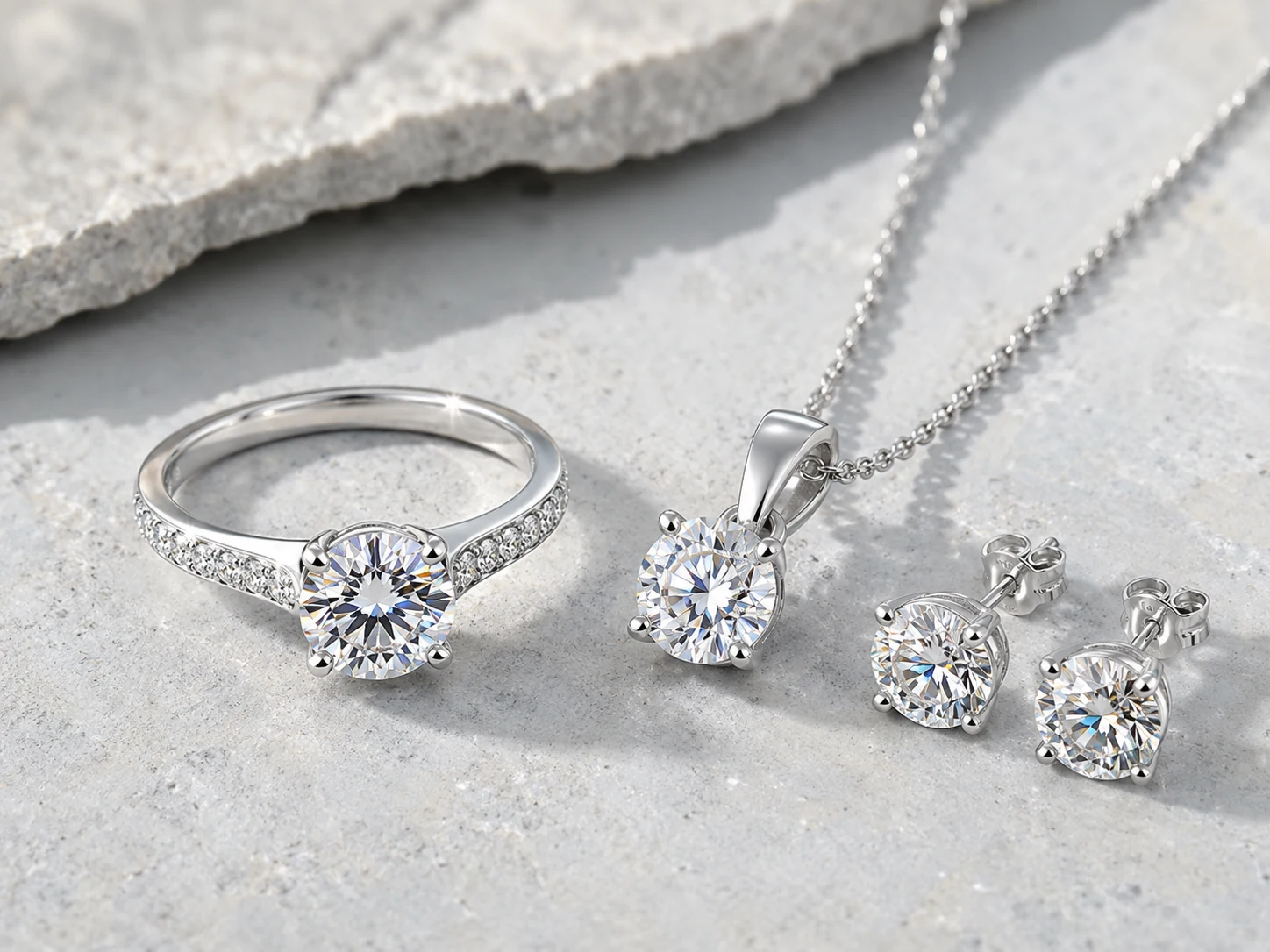 Sterling silver moissanite ring, pendant necklace, and earrings displayed on a cool ivory-gray stone surface.